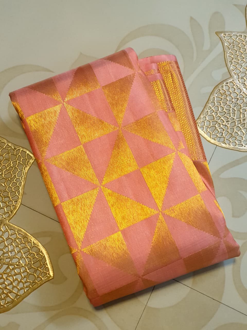 Peach Kanchipuram Handloom 2G Gold Pattu Silk Saree with geometric triangle patterns and a heavy gold border, set against a traditional palace courtyard.