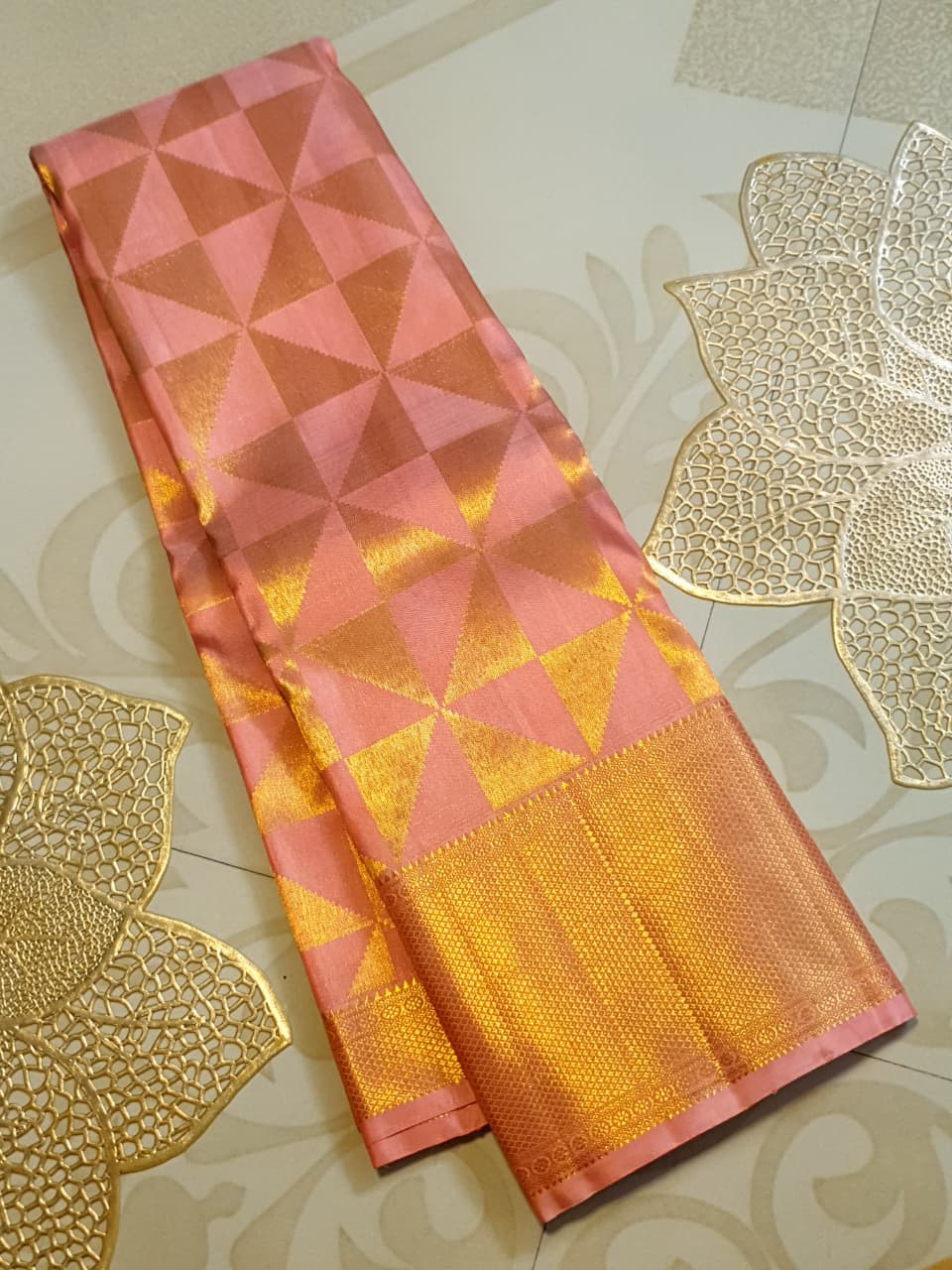 Peach Kanchipuram Handloom 2G Gold Pattu Silk Saree with geometric triangle patterns and a heavy gold border, set against a traditional palace courtyard.