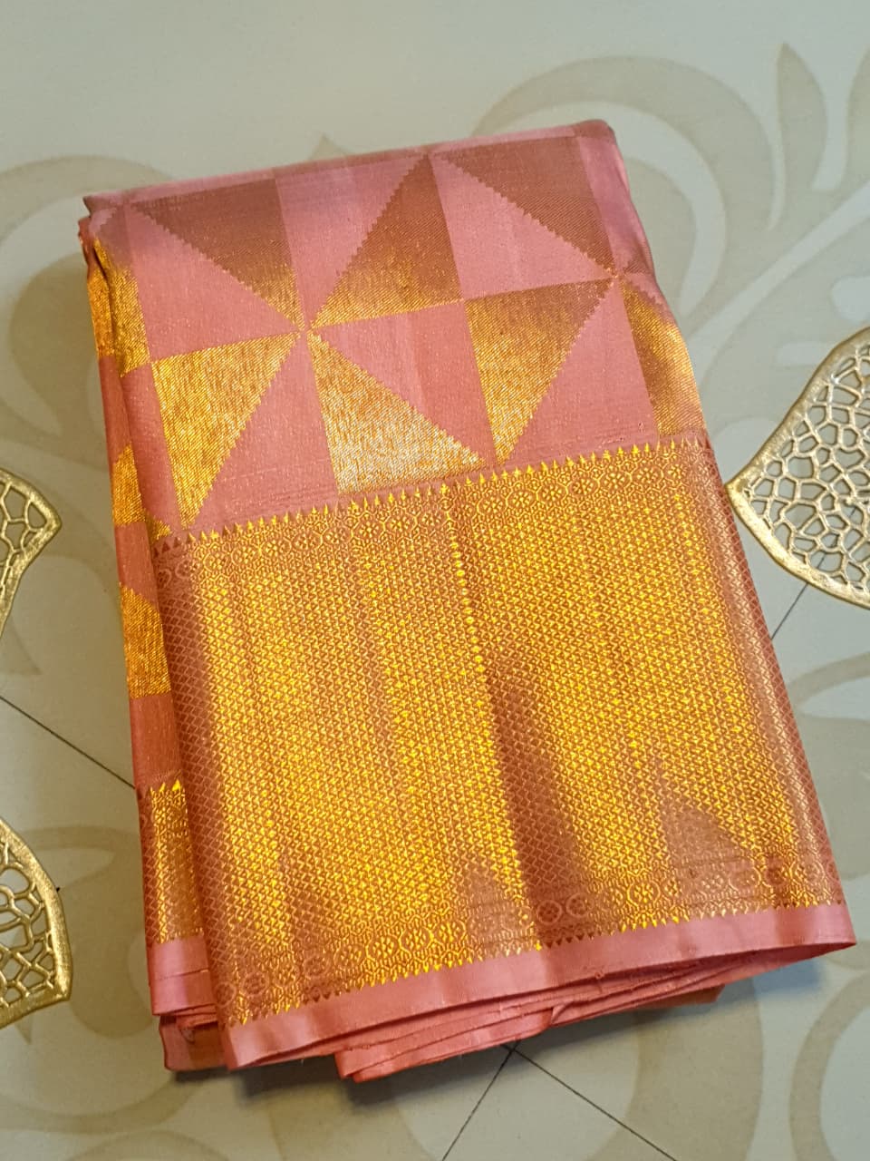 Peach Kanchipuram Handloom 2G Gold Pattu Silk Saree with geometric triangle patterns and a heavy gold border, set against a traditional palace courtyard.