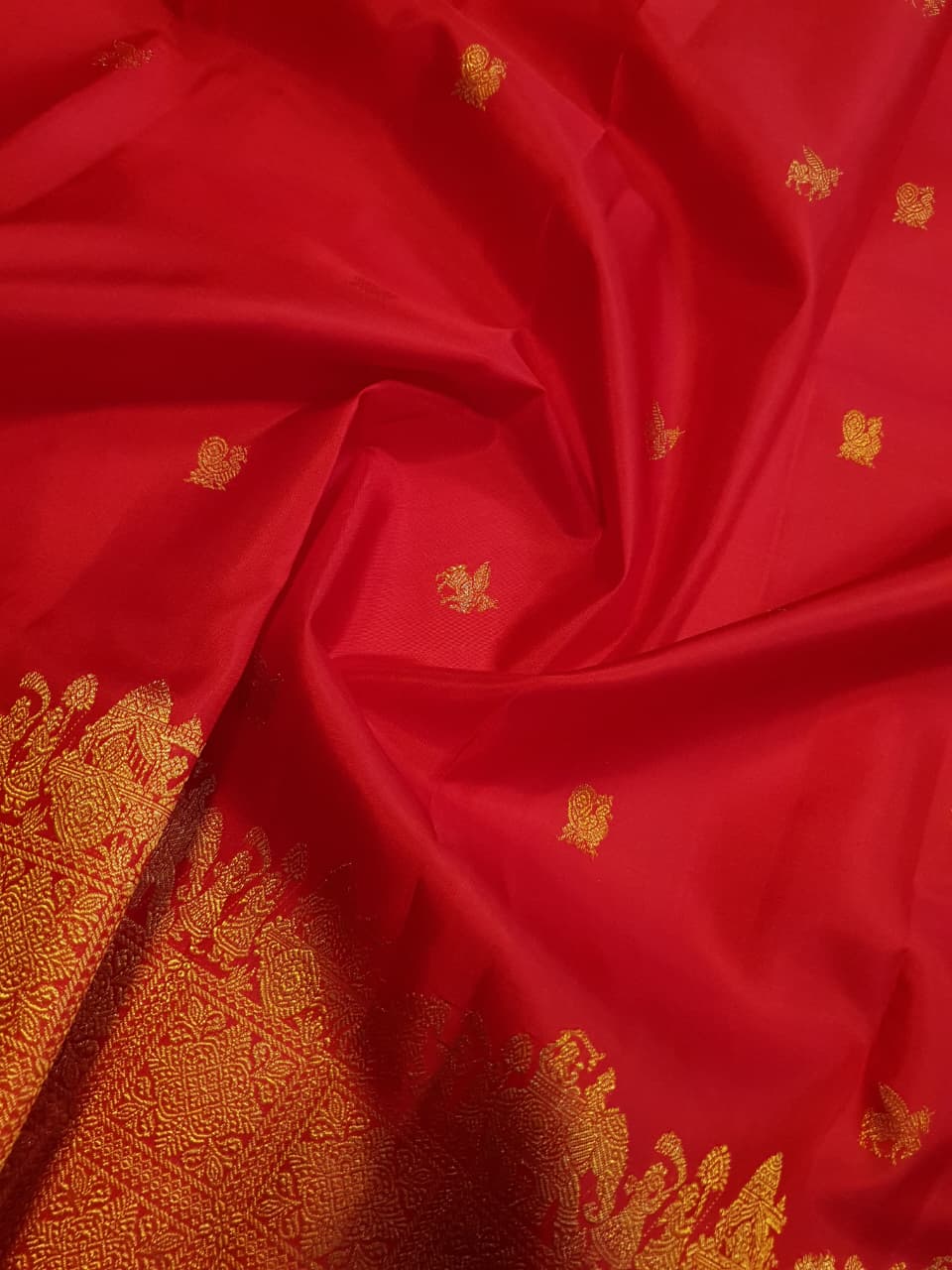A close-up view of a premium 2G Kanjivaram pattu silk saree, highlighting the authentic Silk Mark tag, vibrant red fabric, and detailed gold zari work on the border and body.