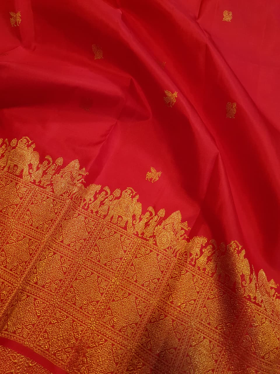 A close-up view of a premium 2G Kanjivaram pattu silk saree, highlighting the authentic Silk Mark tag, vibrant red fabric, and detailed gold zari work on the border and body.