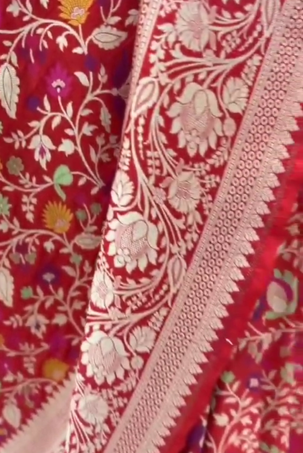 A bright bridal red Banarasi Katan silk saree adorned with colorful Tilfi Meenakari floral buttas and a heavy gold zari border.