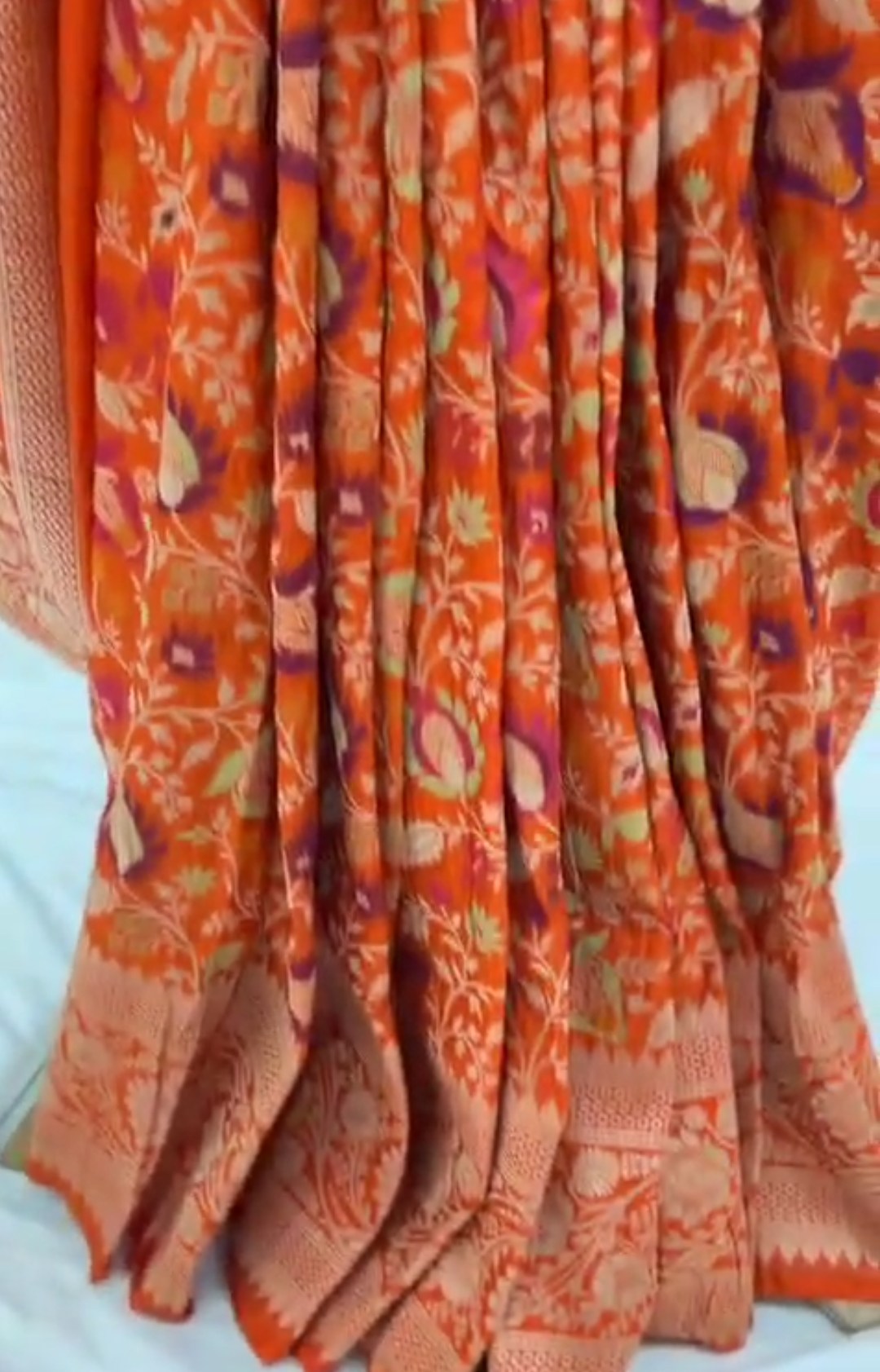 A vibrant orange Banarasi Katan silk saree with multi-colored Tilfi Meenakari floral motifs and an elaborate gold zari border.