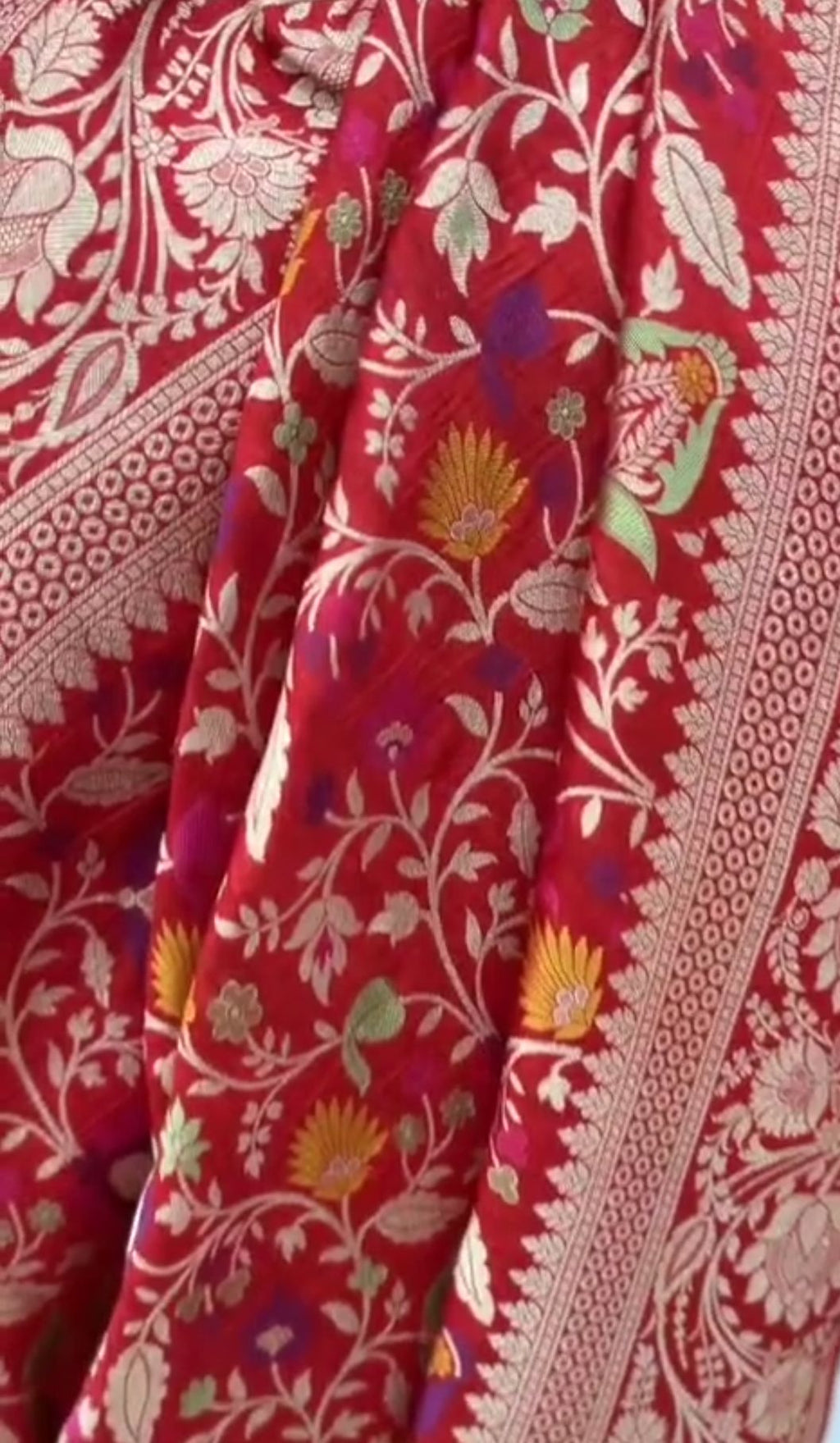 A bright bridal red Banarasi Katan silk saree adorned with colorful Tilfi Meenakari floral buttas and a heavy gold zari border.