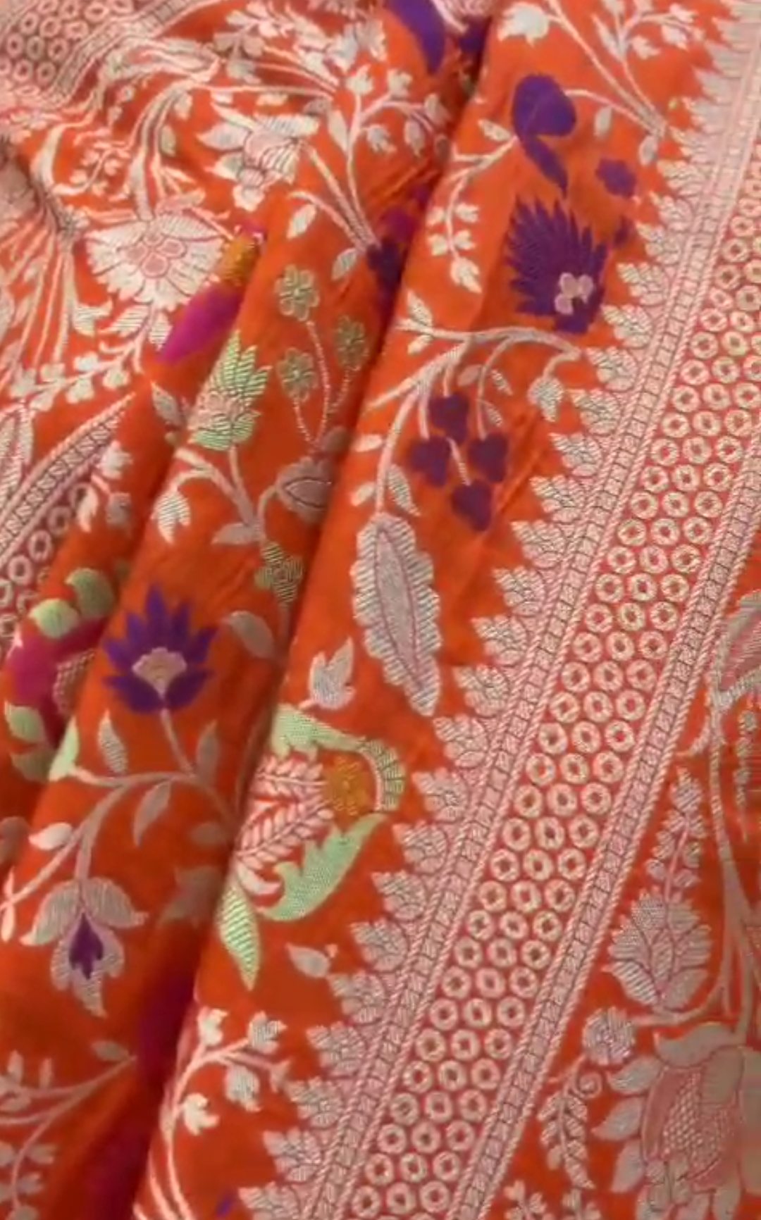 A vibrant orange Banarasi Katan silk saree with multi-colored Tilfi Meenakari floral motifs and an elaborate gold zari border.