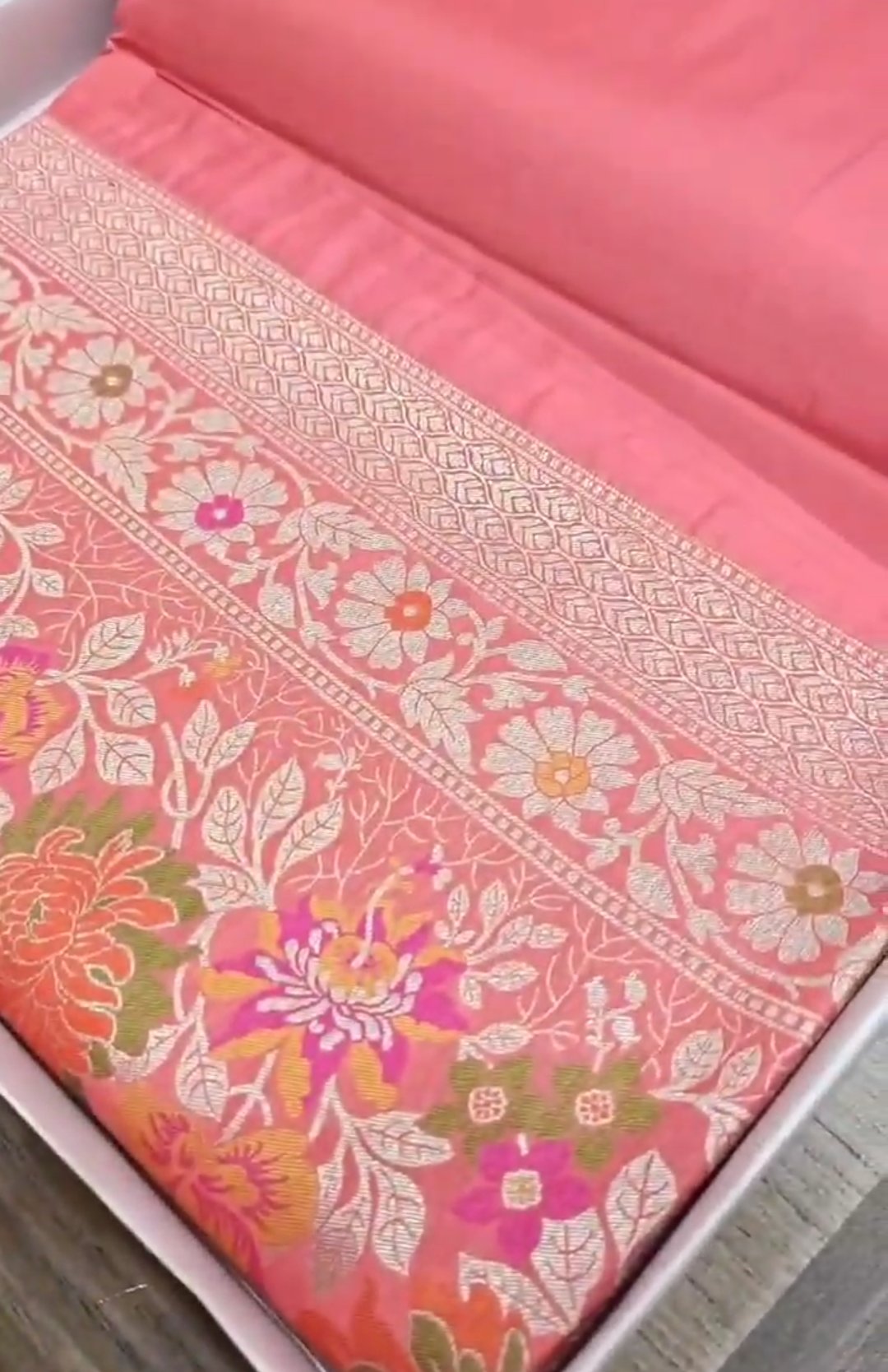 A soft peach Banarasi semi Katan silk saree with a plain body, featuring a multi-colored Tilfi Meenakari floral border and a matching full brocade blouse piece.