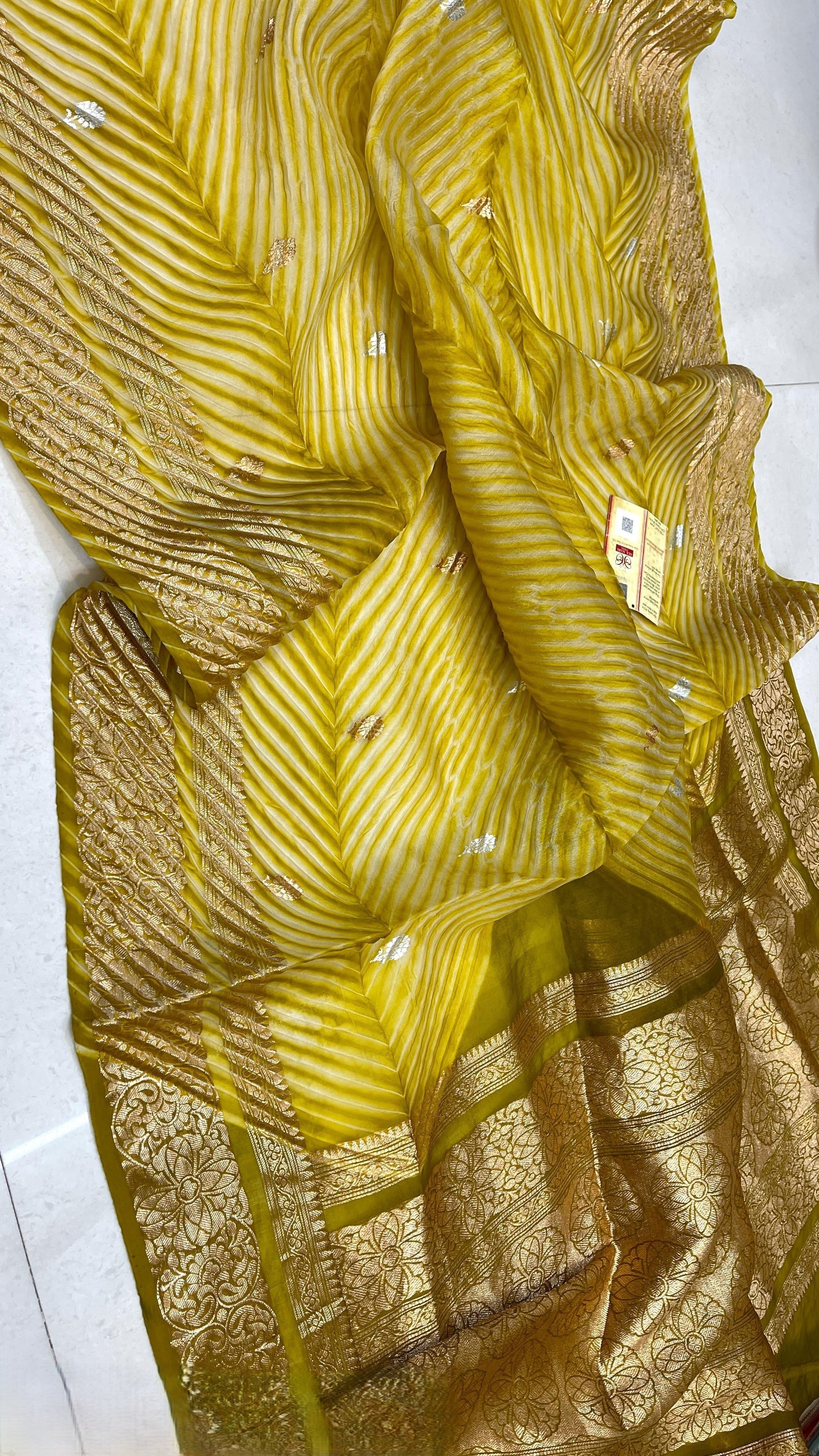 A smiling model in a heritage Banarasi setting wearing a sheer Mustard Yellow Kora Tissue silk saree with diagonal Arasi Shibori pleats and a rich gold zari border.
