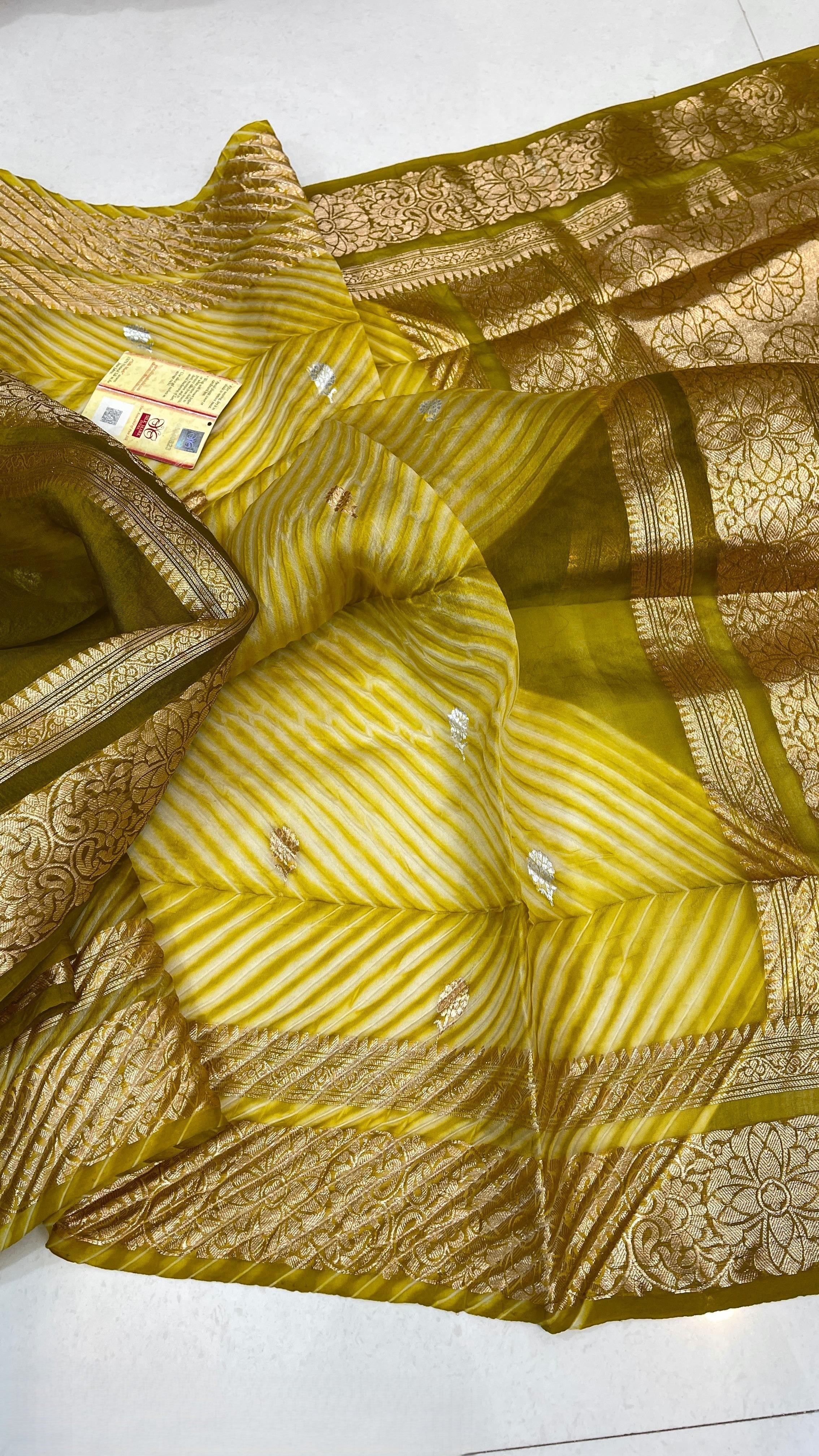 A smiling model in a heritage Banarasi setting wearing a sheer Mustard Yellow Kora Tissue silk saree with diagonal Arasi Shibori pleats and a rich gold zari border.
