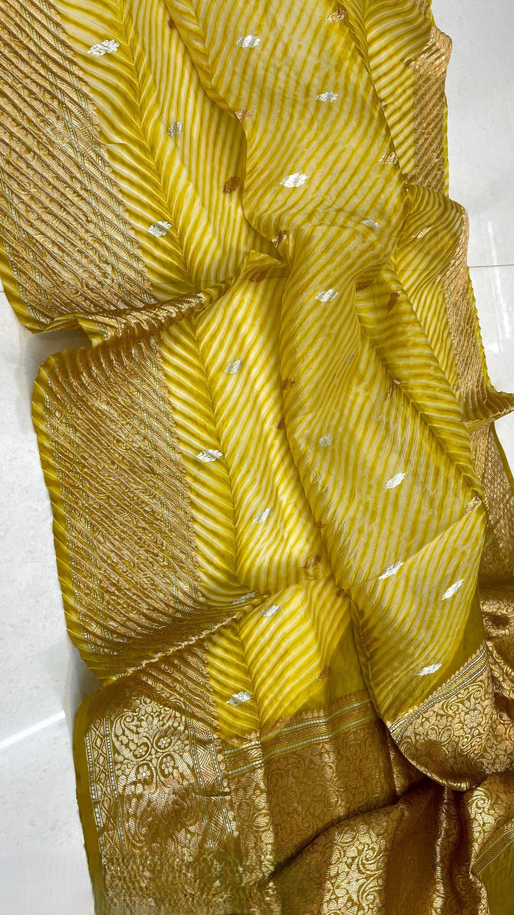 A smiling model in a heritage Banarasi setting wearing a sheer Mustard Yellow Kora Tissue silk saree with diagonal Arasi Shibori pleats and a rich gold zari border.