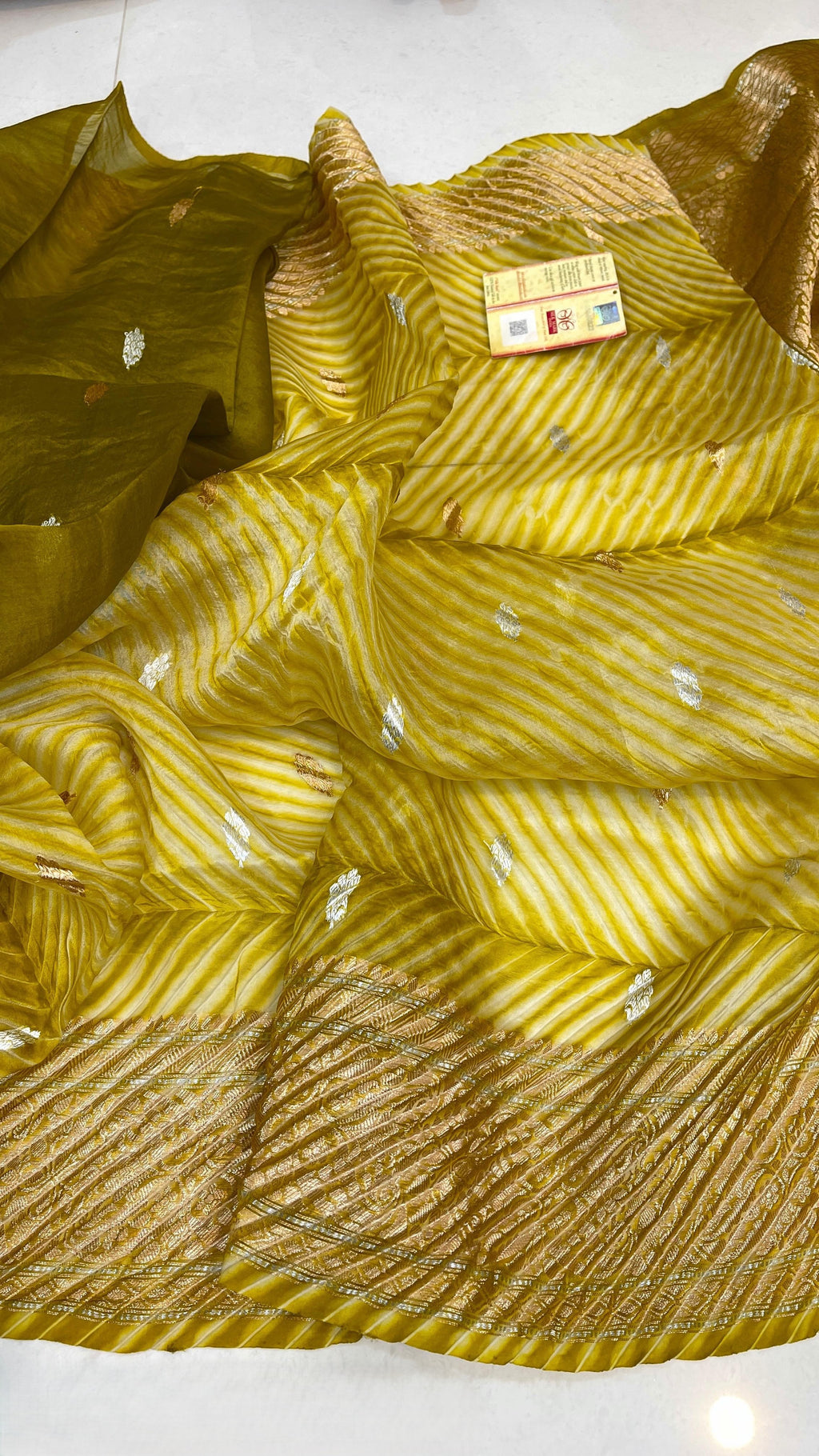 A smiling model in a heritage Banarasi setting wearing a sheer Mustard Yellow Kora Tissue silk saree with diagonal Arasi Shibori pleats and a rich gold zari border.