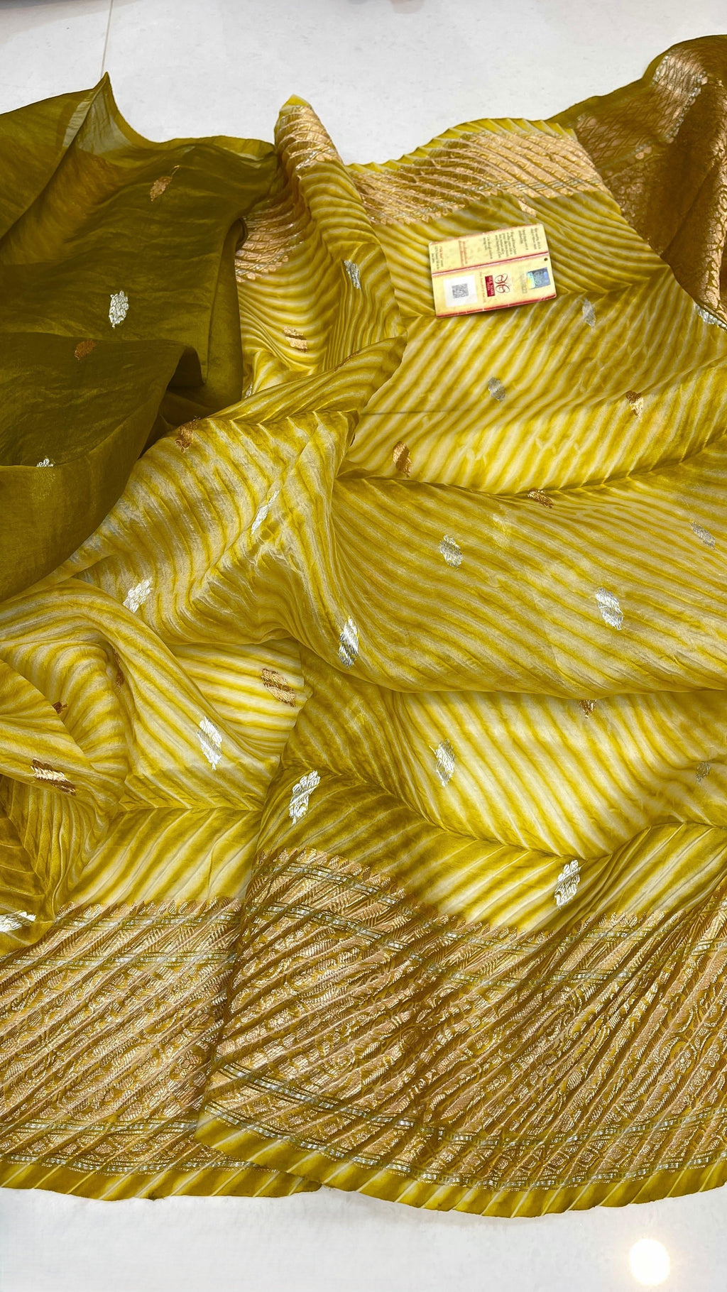 A smiling model in a heritage Banarasi setting wearing a sheer Mustard Yellow Kora Tissue silk saree with diagonal Arasi Shibori pleats and a rich gold zari border.