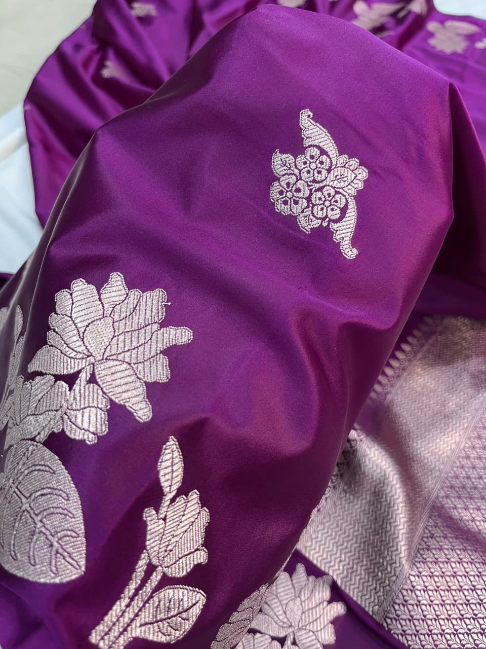 A model presenting a luxurious purple Banarasi Mashru silk saree featuring extensive silver zari floral motifs, a geometric zig-zag border, and a coordinating silver-bordered blouse in a regal heritage setting.