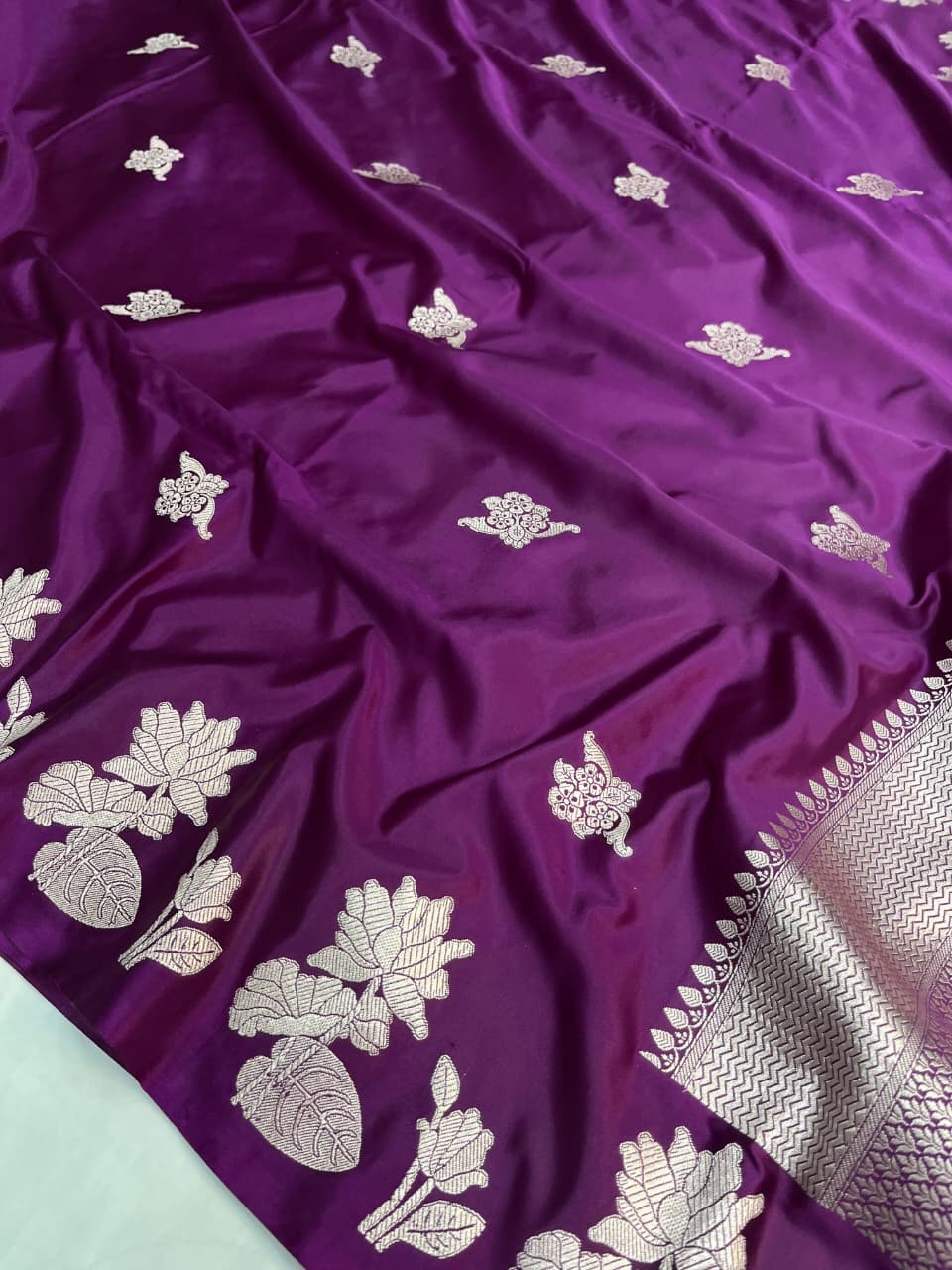 A model presenting a luxurious purple Banarasi Mashru silk saree featuring extensive silver zari floral motifs, a geometric zig-zag border, and a coordinating silver-bordered blouse in a regal heritage setting.