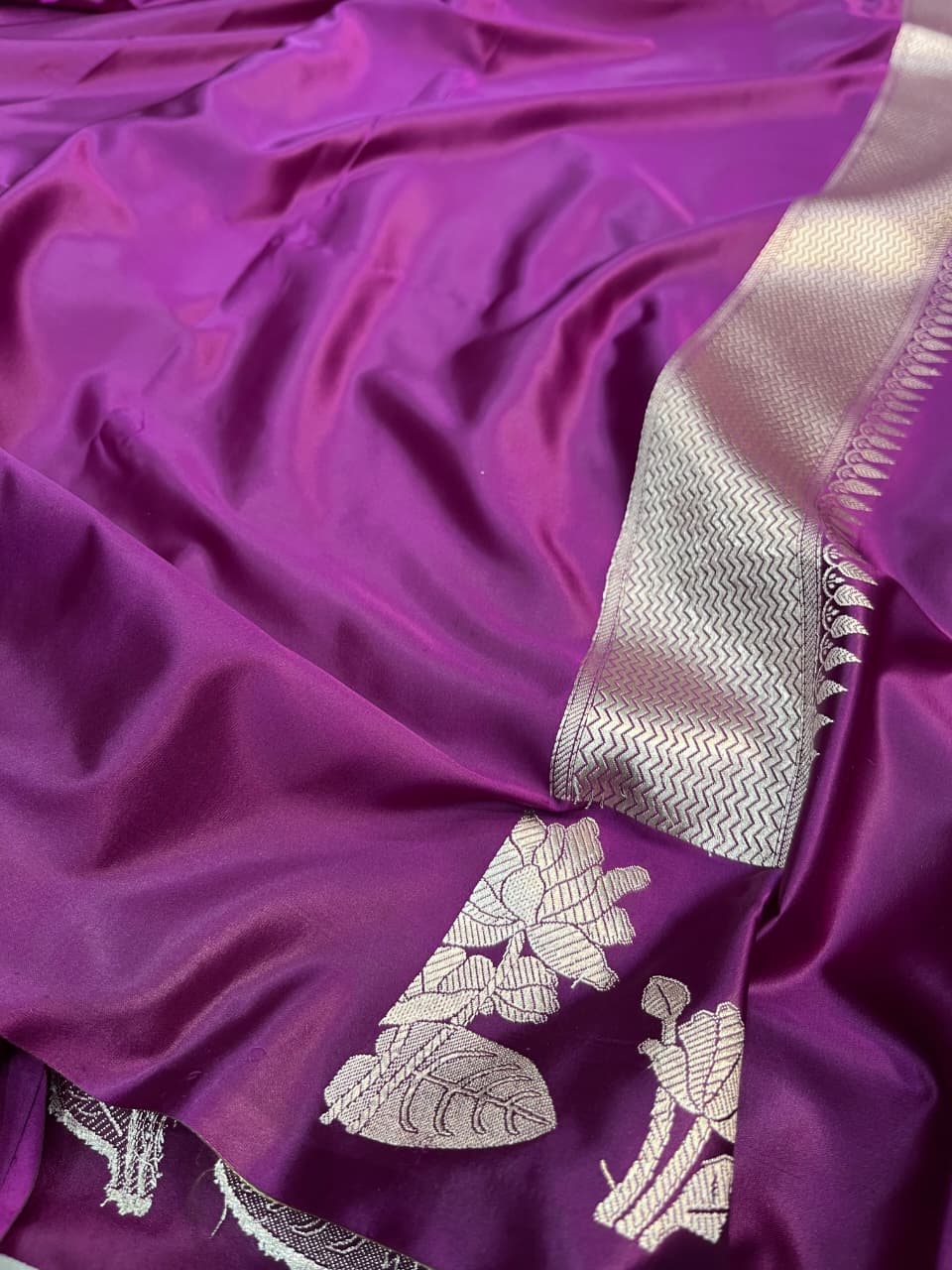 A model presenting a luxurious purple Banarasi Mashru silk saree featuring extensive silver zari floral motifs, a geometric zig-zag border, and a coordinating silver-bordered blouse in a regal heritage setting.