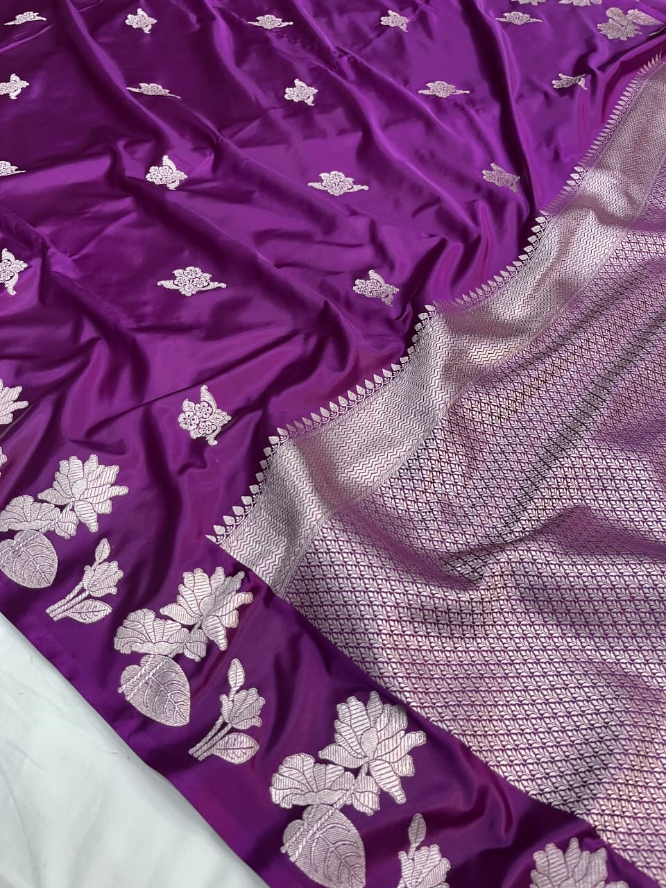 A model presenting a luxurious purple Banarasi Mashru silk saree featuring extensive silver zari floral motifs, a geometric zig-zag border, and a coordinating silver-bordered blouse in a regal heritage setting.