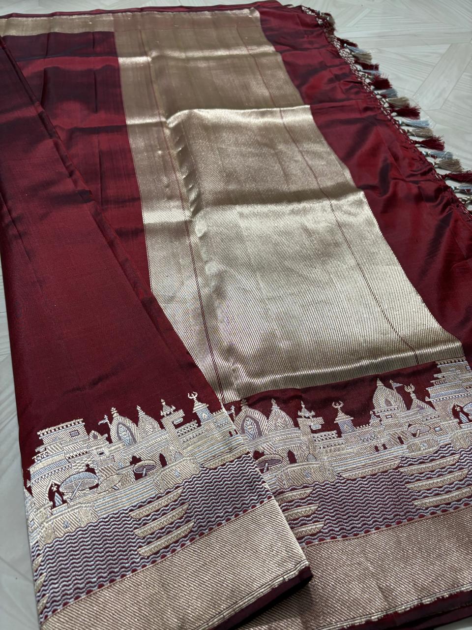 Royal maroon Katan silk saree with a silver and gold zari border depicting the Kashi Ghat temples, worn by a model against a Varanasi riverfront backdrop.