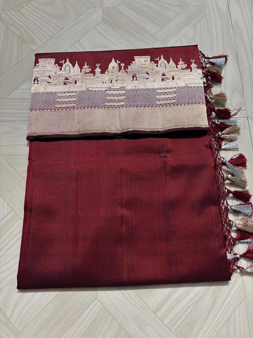 Royal maroon Katan silk saree with a silver and gold zari border depicting the Kashi Ghat temples, worn by a model against a Varanasi riverfront backdrop.