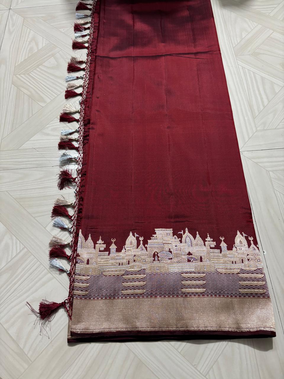 Royal maroon Katan silk saree with a silver and gold zari border depicting the Kashi Ghat temples, worn by a model against a Varanasi riverfront backdrop.