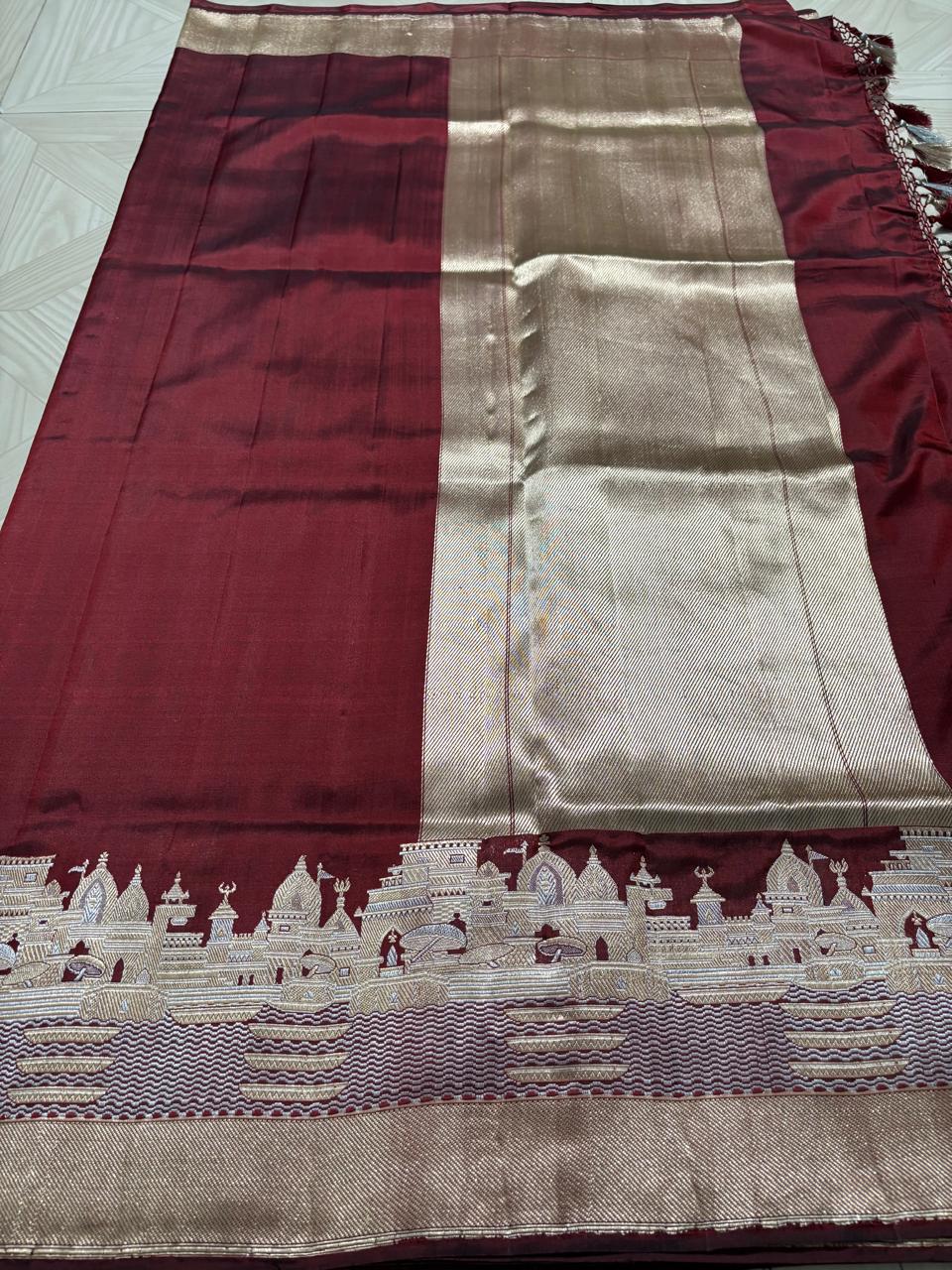 Royal maroon Katan silk saree with a silver and gold zari border depicting the Kashi Ghat temples, worn by a model against a Varanasi riverfront backdrop.
