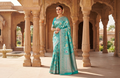 A model gracefully posing in a heritage courtyard wearing a Turquoise Banarasi Handloom Katan Silk Saree with intricate multicolored chakram motifs and ornate zari borders.