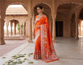 A woman in a traditional setting wears a vibrant orange Banarasi Mashru soft silk saree richly patterned with intricate silver zari work, including floral motifs and a detailed border.