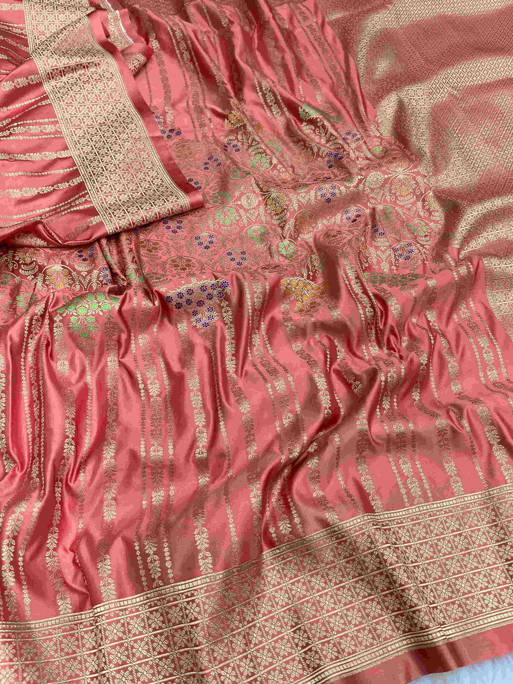 Folded coral pink Banarasi mashru soft silk saree woven with intricate all-over gold zari floral jangla and vibrant meenakari detailing inside a presentation box.