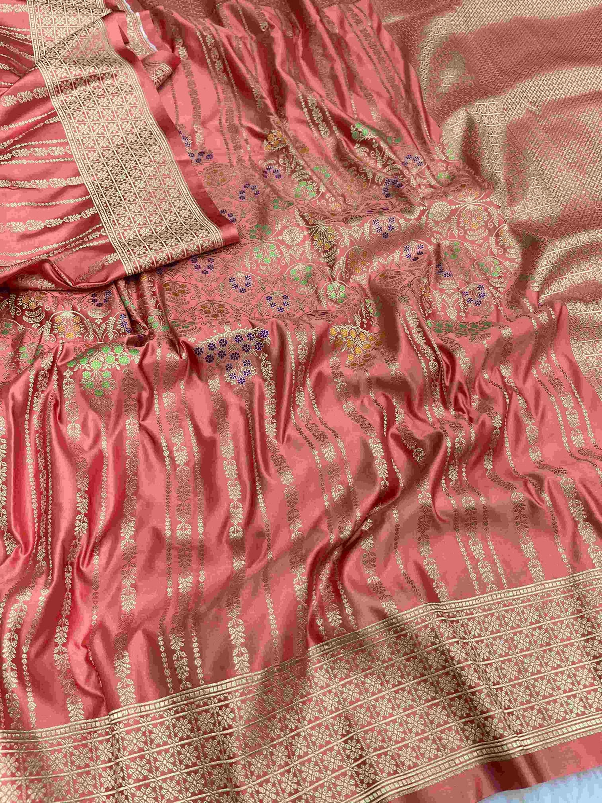 Folded coral pink Banarasi mashru soft silk saree woven with intricate all-over gold zari floral jangla and vibrant meenakari detailing inside a presentation box.