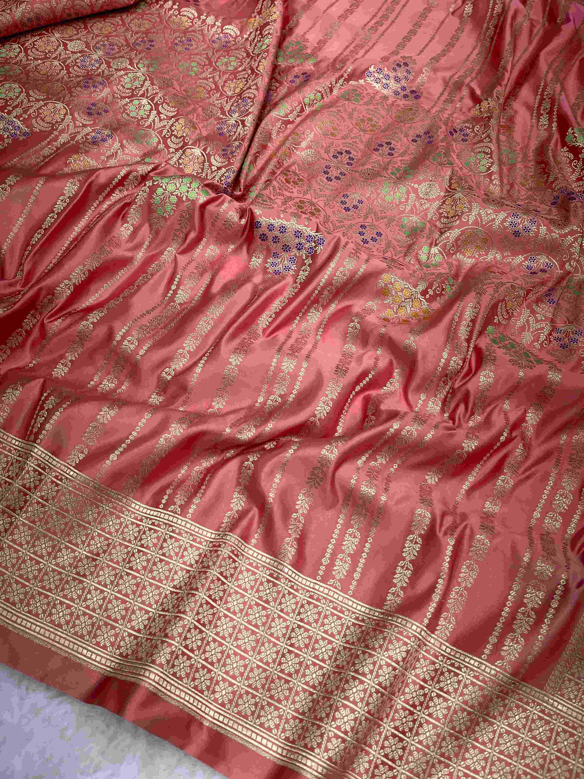 Folded coral pink Banarasi mashru soft silk saree woven with intricate all-over gold zari floral jangla and vibrant meenakari detailing inside a presentation box.