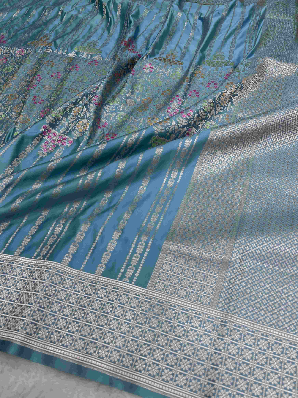Light blue Banarasi mashru soft silk saree featuring all-over golden zari floral jangla weaving with pink, green, and yellow meenakari highlights and a silver geometric border.