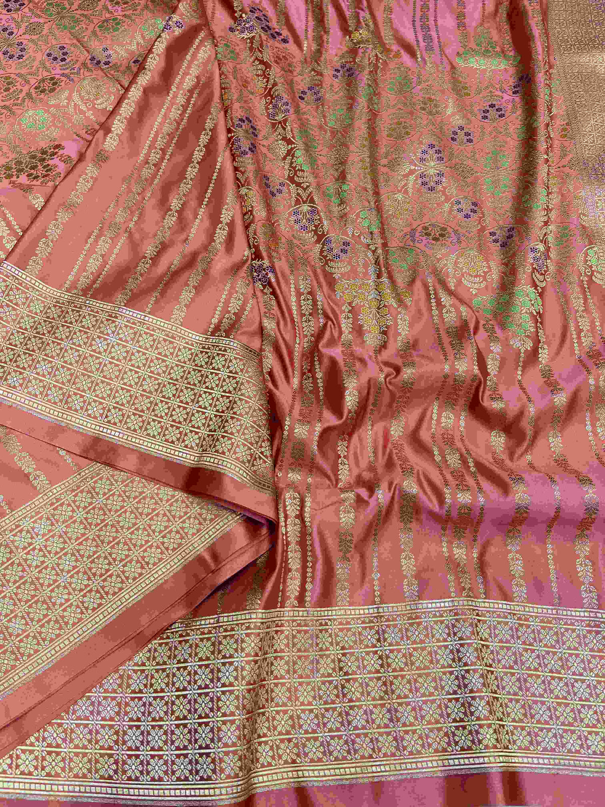 Folded coral pink Banarasi mashru soft silk saree woven with intricate all-over gold zari floral jangla and vibrant meenakari detailing inside a presentation box.