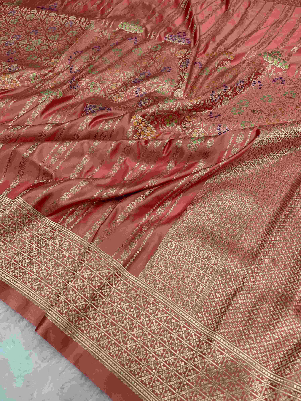 Folded coral pink Banarasi mashru soft silk saree woven with intricate all-over gold zari floral jangla and vibrant meenakari detailing inside a presentation box.