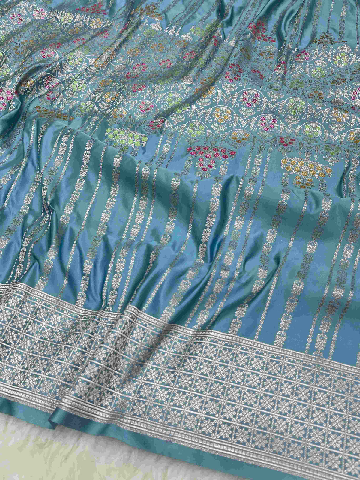 Light blue Banarasi mashru soft silk saree featuring all-over golden zari floral jangla weaving with pink, green, and yellow meenakari highlights and a silver geometric border.