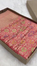 Folded coral pink Banarasi mashru soft silk saree woven with intricate all-over gold zari floral jangla and vibrant meenakari detailing inside a presentation box.