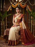 Model wearing an off-white Kanchipuram handloom pattu silk bridal saree with a rich red and gold zari border against a traditional muhuratham background.