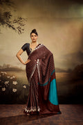 A chocolate brown modal silk saree laid flat, featuring white vertical pinstripes, a large bright teal circle on the pallu, and a matching teal patterned blouse piece.