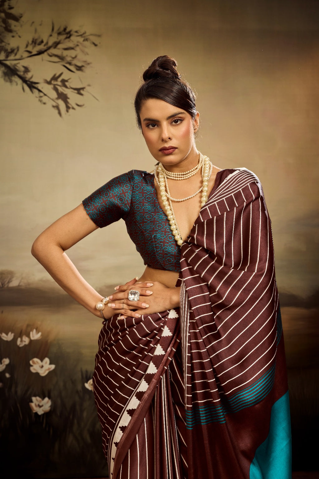 A chocolate brown modal silk saree laid flat, featuring white vertical pinstripes, a large bright teal circle on the pallu, and a matching teal patterned blouse piece.