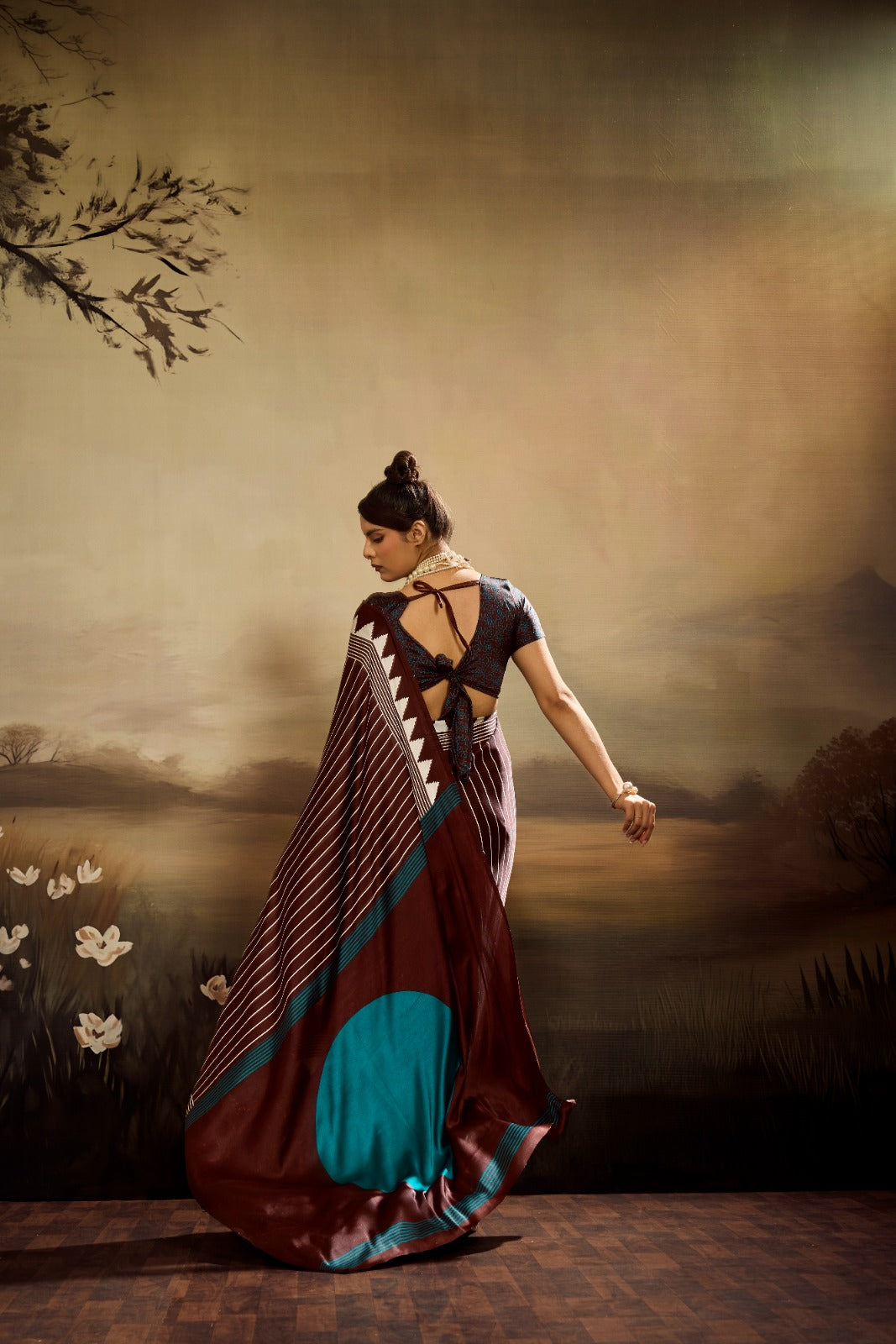 A chocolate brown modal silk saree laid flat, featuring white vertical pinstripes, a large bright teal circle on the pallu, and a matching teal patterned blouse piece.