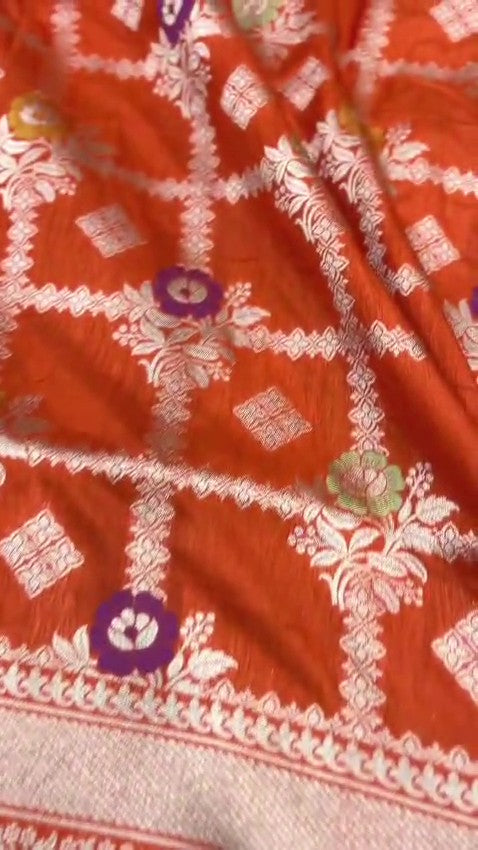 A vibrant orange handloom Banarasi Katan silk saree with an intricate all-over grid pattern and multicolored Meenakari floral motifs, featuring a heavy gold zari border and pallu.