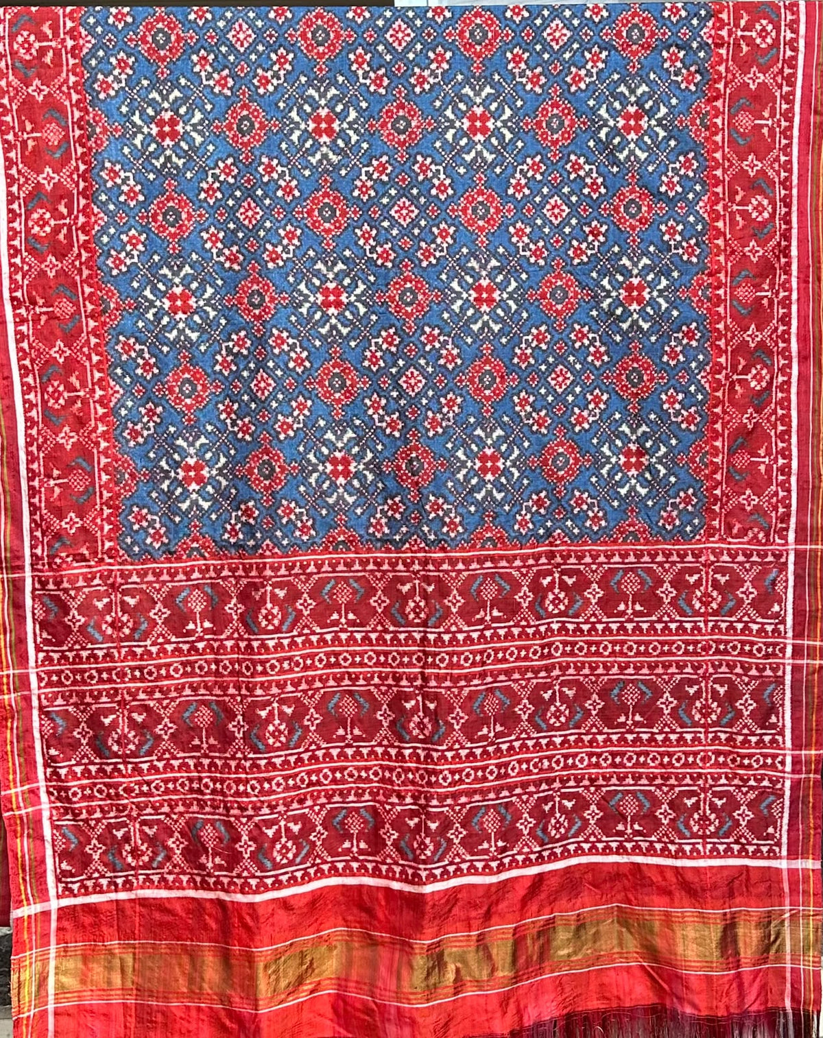 A magnificent blue Patan Patola double ikat pure mulberry silk saree featuring intricate red and white geometric motifs, a striking rich red pallu with traditional tree designs, and contrasting borders.