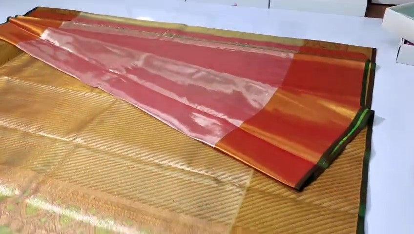 A close-up of a luxurious gold tissue Kanchipuram silk saree with vibrant rust orange borders and intricate Meenakari floral brocade weaving.