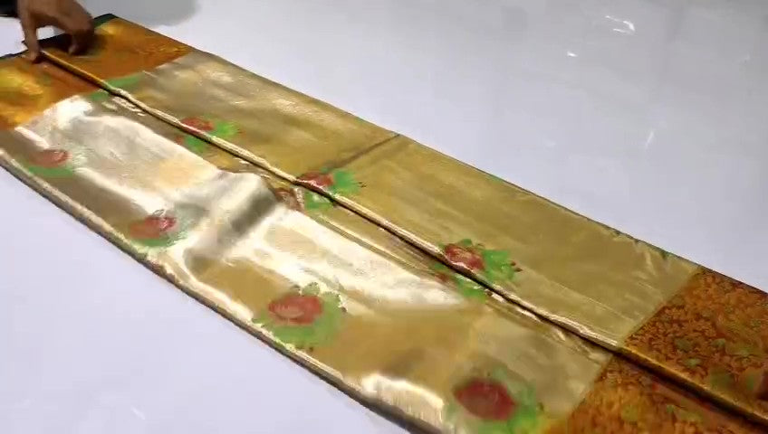 A close-up of a luxurious gold tissue Kanchipuram silk saree with vibrant rust orange borders and intricate Meenakari floral brocade weaving.