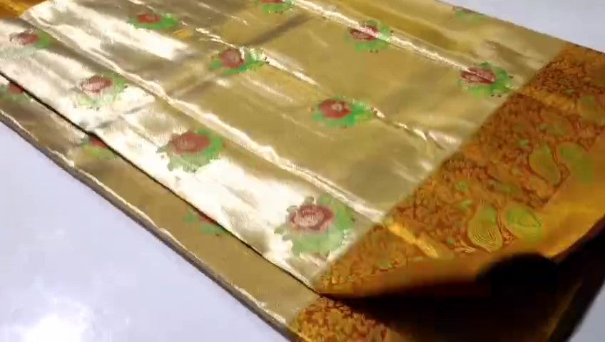 A close-up of a luxurious gold tissue Kanchipuram silk saree with vibrant rust orange borders and intricate Meenakari floral brocade weaving.