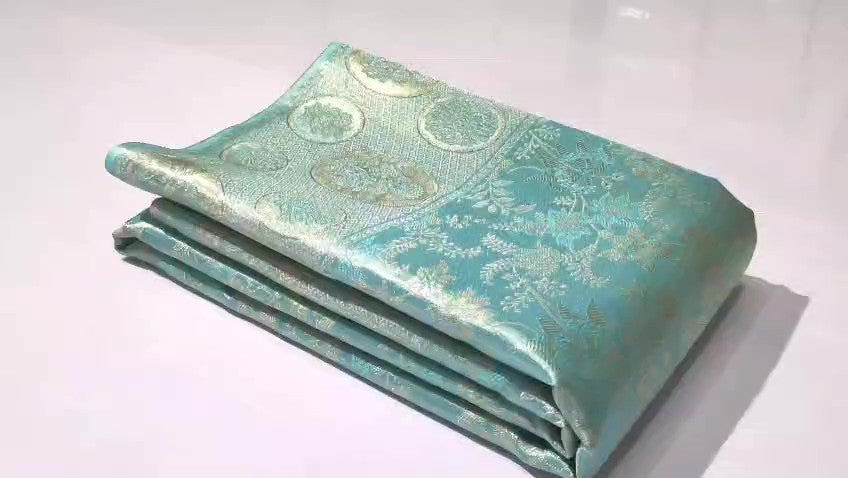 A folded light turquoise brocade fabric with gold floral and circular motifs, displayed on a clean white surface.