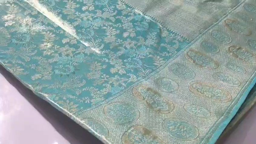 A close-up of a folded turquoise blue brocade fabric with intricate floral and paisley patterns woven in metallic gold and silver threads.