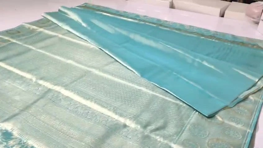 A folded and draped light blue-green silk saree with a shimmering silver border, displayed on a white surface.