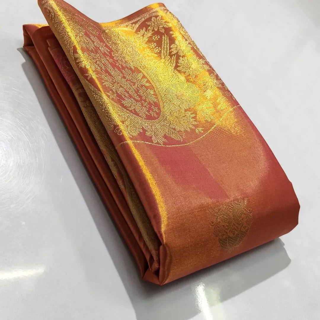 A close-up of a luxurious burnt orange Kanchipuram tissue silk saree with intricate gold brocade weaving, authentic Silk Mark certification, and a shimmering metallic finish.