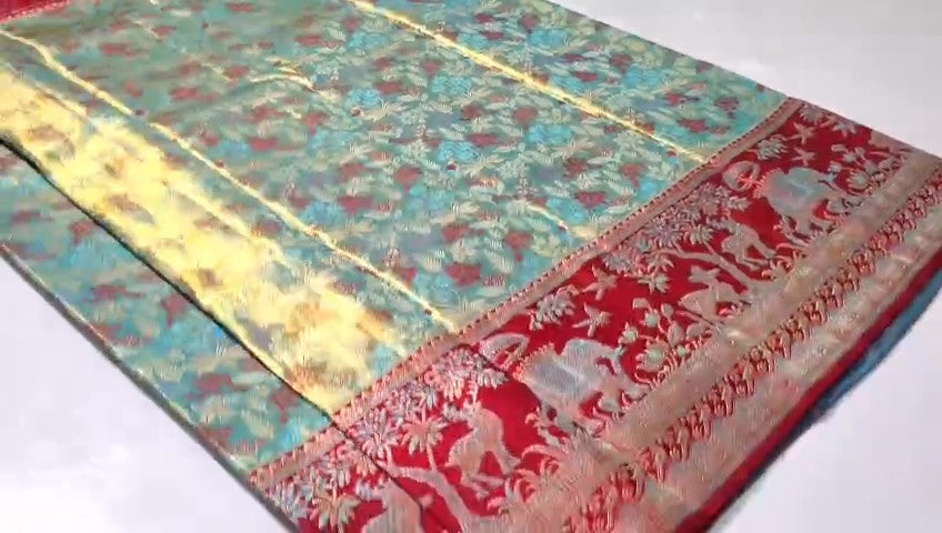 A close-up of a traditional woven textile, likely a sari, featuring a turquoise blue body with red floral motifs, a wide yellow-gold border, and a red border with white zari animal and floral patterns.